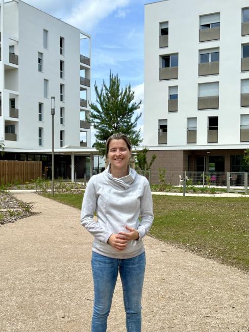 une femme blonde dans un jardin avec un immeuble blanc en fond 